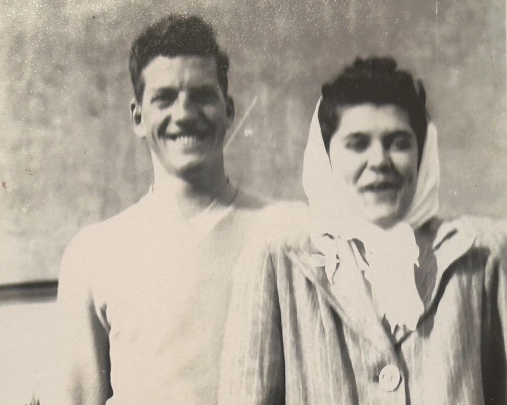 A man in a light shirt stands next to a woman wearing a headscarf and a striped coat. Both are smiling at the camera. The black and white photo captures them on their family farm.